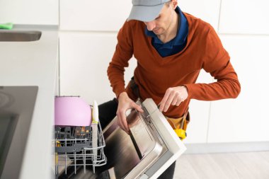 Ev mutfağındaki bulaşık makinesinin tamiri sırasında işçi kıyafeti giymiş genç modern bir tamirci..