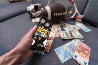 Çevrimiçi spor bahsi. Akıllı telefon ve futbol topu olan bir elin arka planına dolar düşüyor. Yaratıcı altyapı, kumar