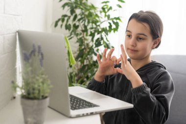 Distant education. Friendly teen girl having online lesson via laptop and showing gesturing in sign language, studying online from home.