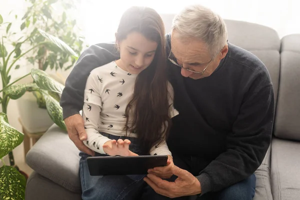 귀여운 소녀와 잘생긴 할아버지는 집에서 소파에 미소짓고 태블릿을 — 스톡 사진