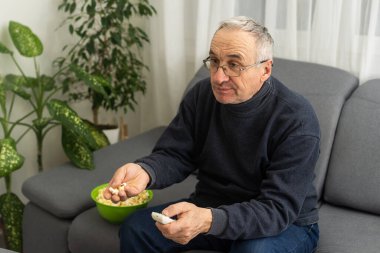 Portrait of elderly man watching TV with popcorn and a remote control.