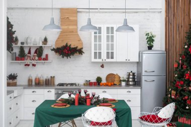 Çam çelenkleri ve Noel oyuncakları ile süslenmiş bir mutfak. Masa ve yakındaki Noel ağacı kadar değerli. Noel için dekore edilmiş parlak bir mutfak.