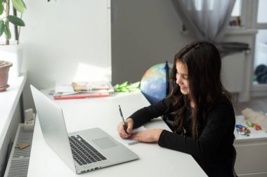 Evde eğitim. Bir kız, bir öğretmen ve sınıf ile okul dersinin online görüntülü sohbeti sırasında dizüstü bilgisayarla masada oturuyor. Uzaktan eğitim kavramı. Karantinada kendini izole etme