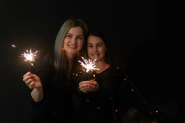 Happy family in black - woman and little girl, with a sparkler in hand the New Years Christmas at home. The girl at the father on hands. New year 2023