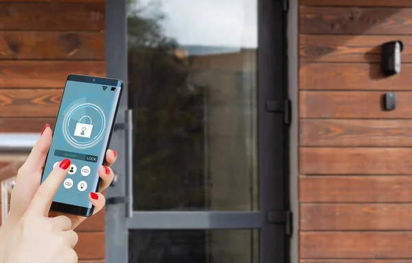 Female hand holding black mobile smart phone with smart home application on the screen. Blurred house on the background. For access to all of the controls of your house and caring of home security.