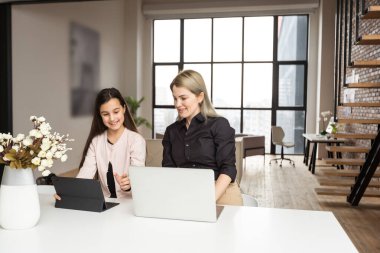 Portrait of mother and daughter sitting in living room. Cute girl holding in her hand a digital tablet while her mom using laptop and looking at something together