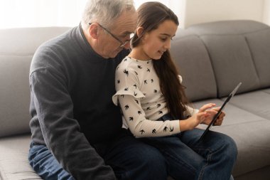 Happy family senior grandfather and girl watching cartoon on tablet on weekend day at home together.