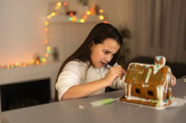 Çocuklar Noel kurabiyesi evi pişiriyor. Çocuklar kış tatilini evde kutluyorlar. Şömine ve ağaçla dekore edilmiş oturma odası. Aile aktivitesi. Küçük kız kurabiye yapıyor..