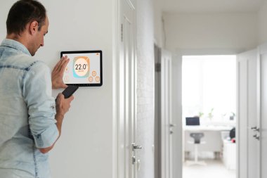 Genç adam oturma odasındaki sıcaklığı akıllı telefon ve dijital dokunmatik ekranlı panelle kontrol ediyor. Akıllı bir evde ısıtma kontrolü kavramı. Yüksek kalite fotoğraf