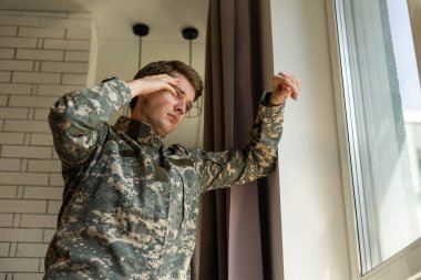 Serious short-haired young man in military uniform soldier looking through the window, upset soldier suffering from posttraumatic syndrome after returning home, copy space, closeup