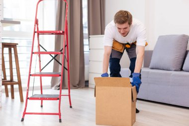 Depoda rafların ve rafların arasında çalışan karton bir paket tutan genç adam. Kutulu teslimatçı. Personel işçisi, turuncu üniforma şapkası, tişört, tulum servisi siparişleri getiriyor..