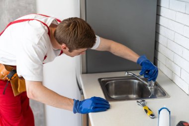 Erkek tesisatçı müşteri evinde lavabo musluğu tamir ediyor. Tamirci ya da tamirci müşteri mutfağında boru hattını tamir eder. Kaliteli tesisat şirketi servisi. Bakım ve ev yardımı kavramı.