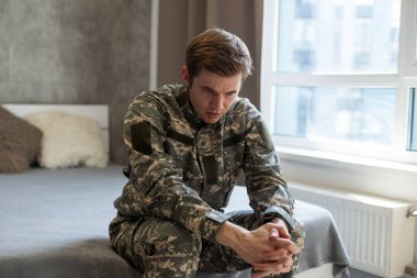 Depresif düşünce ve anılara dalmış stresli bir asker. Üzücü, kamuflaj üniformalı genç bir asker koltukta oturup savaşı düşünüyor. Travma Sonrası Stres ve Terapi