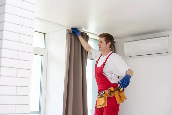 A male repairman installs a curtain rod and hangs curtains in the house. services to help with household chores and repairs.