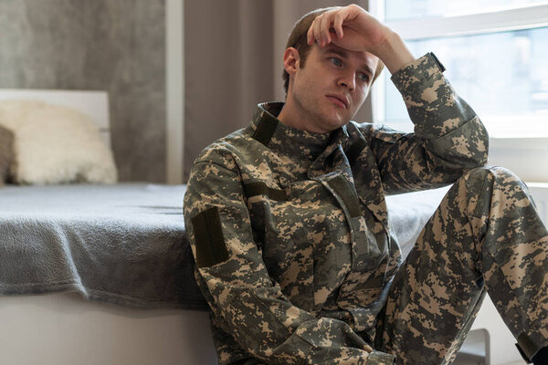 Stressed veteran soldier engrossed in depressive thoughts and memories. Sad depressed young military man in camouflage uniform sitting on couch and thinking about war. PTSD and therapy concept