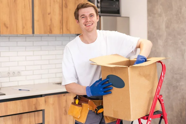 stock image Smiling male worker of moving and delivery company holding cardboard box. Loader in overalls posing against background of colleague who packs cardboard boxes. Moving service concept