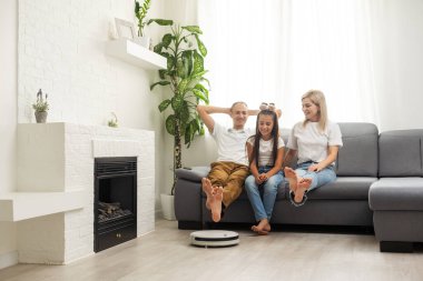 Young family resting on the couch while robotic vacuum cleaner doing its work at home. Household robots concept