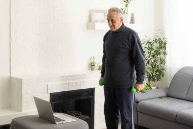 Sport in mature age. Happy middle-aged man doing stretching exercises in front of laptop at home, watching online tutorials, in living room interior, free space.