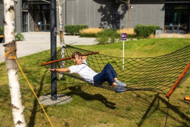 Mutlu neşeli çocuk gülümseyen ve yalan söyleyen, ormandaki bir hamakta dinlenen açık yeşil çimenlerin arasında. Yüksek kalite fotoğraf