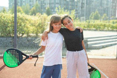Çocuklar ve spor konsepti. Padel kortunda raketler ve tenis toplarıyla poz veren gülümseyen kızların portresi. Yüksek kalite fotoğraf