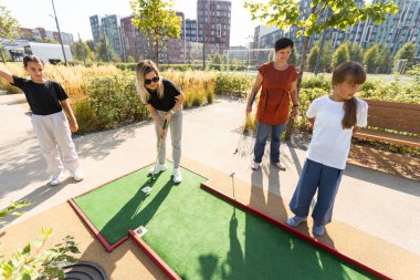Ailesiyle mini golf oynayan tatlı bir kız. Yeni yürümeye başlayan çocuk dışarıda eğleniyor. Çocuklar ve yetişkinler için açık havada yaz sporu. Aile tatilleri ya da tatil köyleri. Yüksek kalite fotoğraf