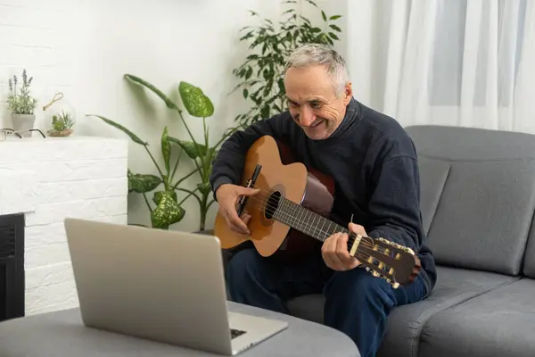 Portrait of senior man in headphones taking online guitar lesson looking at laptop screen. Retired male learning to play guitar watching webinar on computer at home.