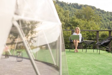 Serbest çalışan genç bir kadın, sıcak ve güneşli bir havada, dağların yakınındaki açık havada, balkonda uzaktan kumandalı bir dizüstü bilgisayar üzerinde çalışıyor.