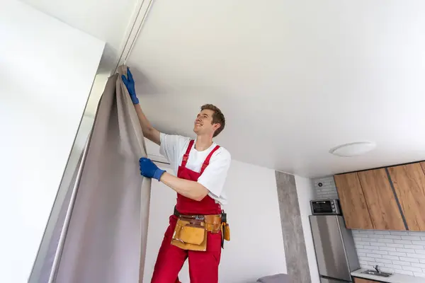 stock image A male repairman installs a curtain rod and hangs curtains in the house. services to help with household chores and repairs.