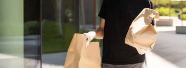 Koruyucu maskeli ve eldivenli teslimatçı. Bir binanın önünde poşet dolusu erzak tutuyor.