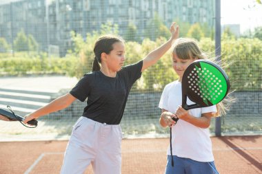 Raket ve topları olan genç kızlar Padel sahasında kameraya bakıp gülümsüyorlar. Yüksek kalite fotoğraf