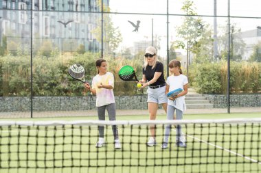 Aktivite giyimli genç bir tenis antrenörü, stadyum eğitimi sırasında iki tatlı kızdan birinin elinde raket tutuyor. Yüksek kalite fotoğraf