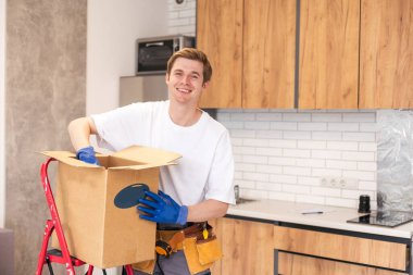 Depoda rafların ve rafların arasında çalışan karton bir paket tutan genç adam. Kutulu teslimatçı. Personel işçisi, turuncu üniforma şapkası, tişört, tulum servisi siparişleri getiriyor..