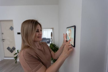 Oturma odasındaki duvara dijital bir dokunmatik ekranla evi kontrol eden kadın..