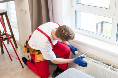 Genç, yakışıklı, profesyonel tesisatçı yeni inşa edilmiş bir evin boş bir odasına ısıtma kaloriferi yerleştiriyor. İnşaat, bakım ve onarım konsepti.