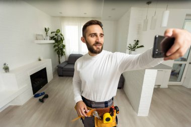Yakışıklı, neşeli tamircinin portresi. Akıllı telefondan selfie çekiyor. Gömlek ve tulum giyiyor..