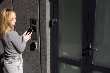 Bir kadın akıllı telefon kullanarak giriş kapısına akıllı kilit takıyor. Anahtarsız erişime sahip akıllı elektronik kilitler kullanma kavramı.