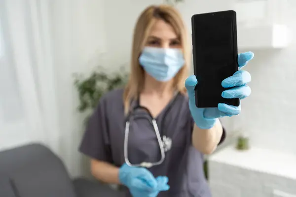 Young woman in the nurse uniform with stethoscope and wearing protective facemask point her finger to smartphone to show Covid-19 vaccinated digital certificate application with copy space