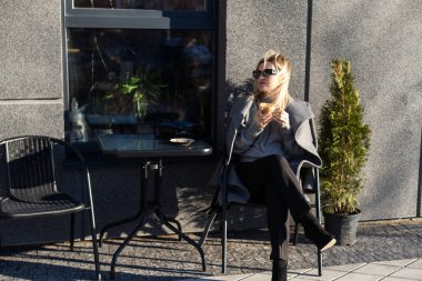 Kahve içen genç bir kadın, güneşli bir günde şehrin eski caddesindeki terasta cep telefonuyla oturuyor. Yüksek kalite fotoğraf