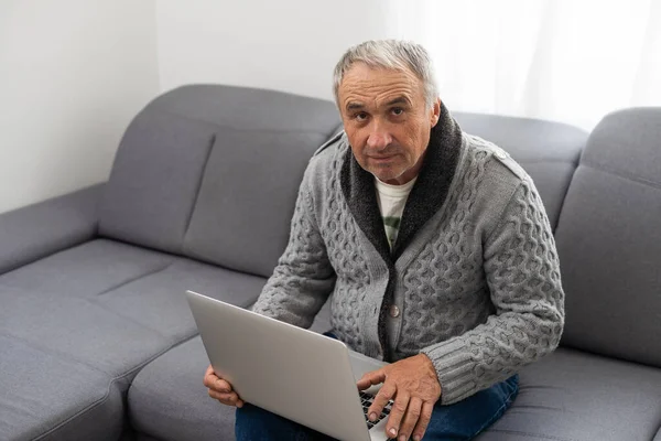 Older man sitting on sofa, smiling at computer screen at home. High quality photo