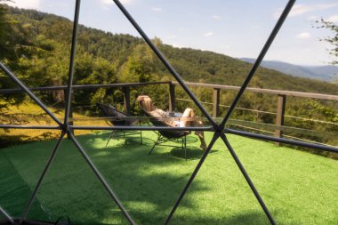 Genç bir kadın, modern evin yakınındaki ahşap terasta, çam ormanının yakınındaki panoramik pencereli güneş yatağında dinleniyor. Yalnızlık ve doğada eğlence kavramı.