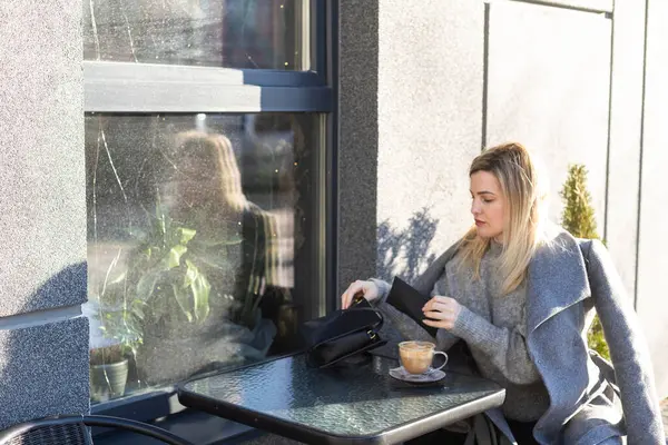 Kahve içen genç bir kadın, güneşli bir günde şehrin eski caddesindeki terasta cep telefonuyla oturuyor. Yüksek kalite fotoğraf