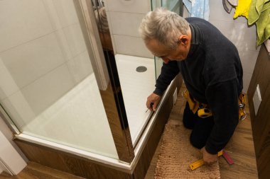 an elderly man repairing door of shower cabin in bathroom.