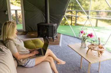 Kubbe kampında dizüstü bilgisayar kullanarak internette sohbet eden mutlu genç bir kadın. Glamping tatil konsepti.