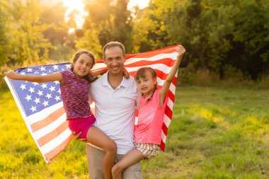 Gün batımında buğday tarlasında Amerikan bayrağı taşıyan mutlu bir aile. Bağımsızlık Günü, 4 Temmuz
