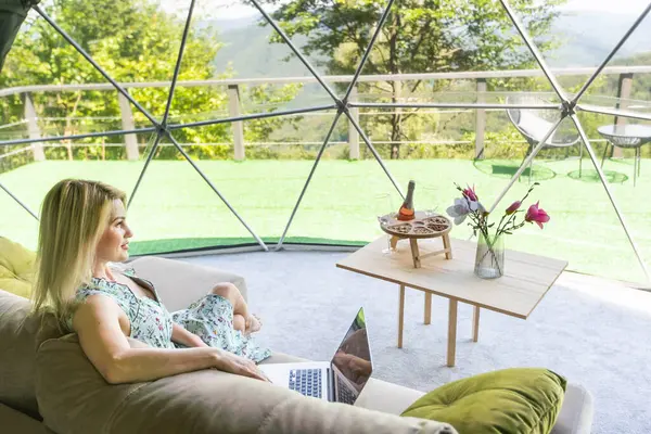 stock image Transparent bubble tent at glamping, Lush forest around and interior. Woman with laptop.