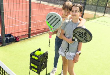 Çocuklar ve spor konsepti. Padel kortunda raketler ve tenis toplarıyla poz veren gülümseyen kızların portresi. Yüksek kalite fotoğraf