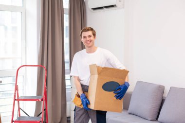 Depoda rafların ve rafların arasında çalışan karton bir paket tutan genç adam. Kutulu teslimatçı. Personel işçisi, turuncu üniforma şapkası, tişört, tulum servisi siparişleri getiriyor..