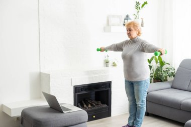 Olgunluk çağında spor. Mutlu son sınıf kadını evde dizüstü bilgisayarın önünde egzersiz yapıyor, online dersler izliyor, boş yer arıyor.