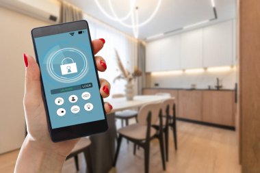 Elinde akıllı telefon tutan ve evinde güvenlik uygulaması olan genç bir kadın. Mobil bir cihazdan ev güvenliğini kontrol etme ve yönetme kavramı. Yüksek kalite fotoğraf