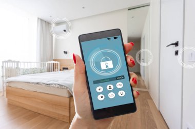 Elinde akıllı telefon tutan ve evinde güvenlik uygulaması olan genç bir kadın. Ev güvenliğini mobil bir cihazdan kontrol etme ve yönetme kavramı.